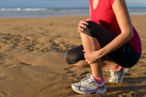 Female runner clutching her shin because of a running injury and inflammation. Tibial periostitis hurt while jogging on beach.